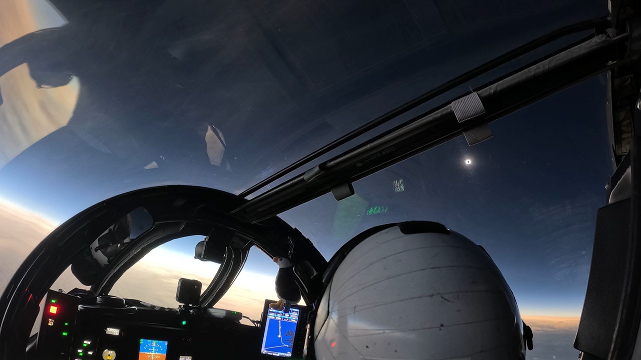 NASA-videostill vanuit de cockpit: de verduisterde zon boven een donkere hemel, met een lichte horizon en een helm op de voorgrond.