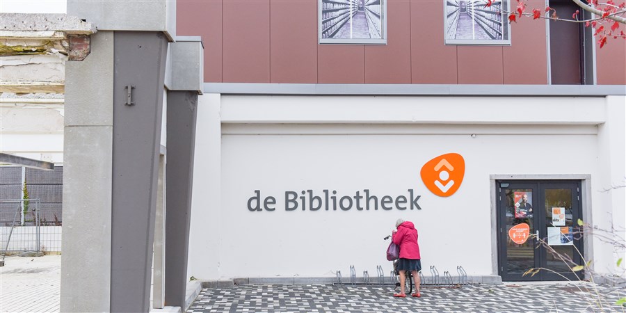 Originele CBS-foto van de ingang van een openbare bibliotheek, met fietsenrekken en een bezoeker bij de gevel.